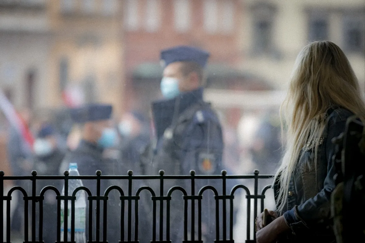 Woman standing by a fence with blurred police officers beyond it